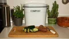 Keuken compostbak met groenteafval op snijplank en verse kruiden op achtergrond.